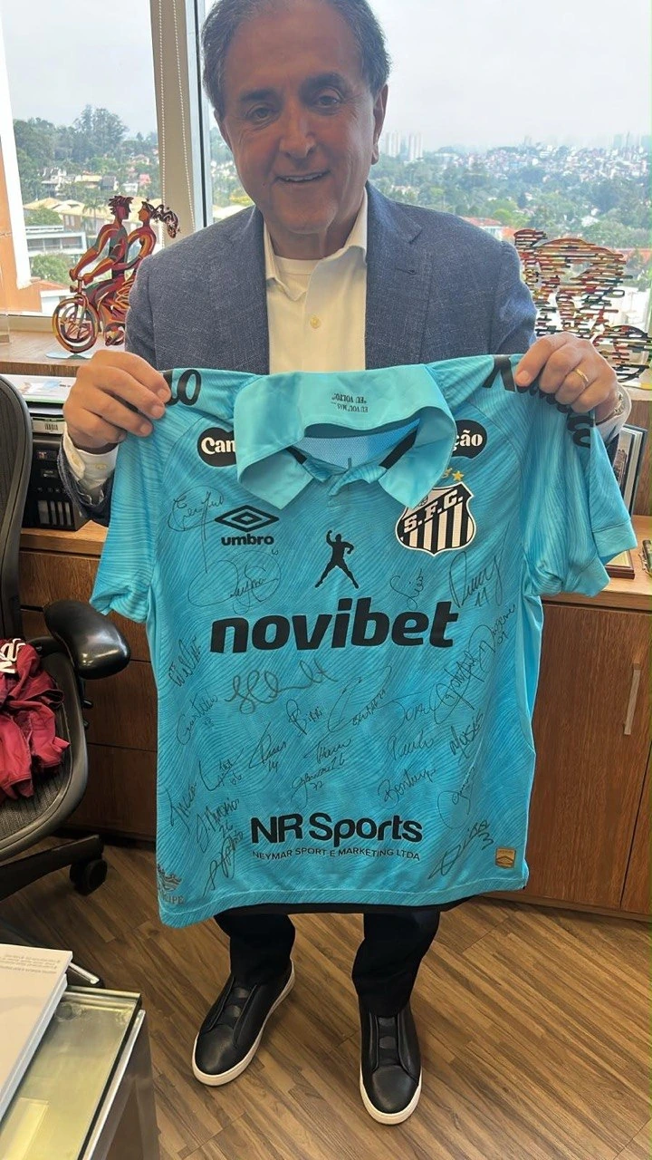 Pessoa em ambiente interno apresentando camisa turquesa do Santos com assinaturas, escudo do clube e logotipos de patrocinadores